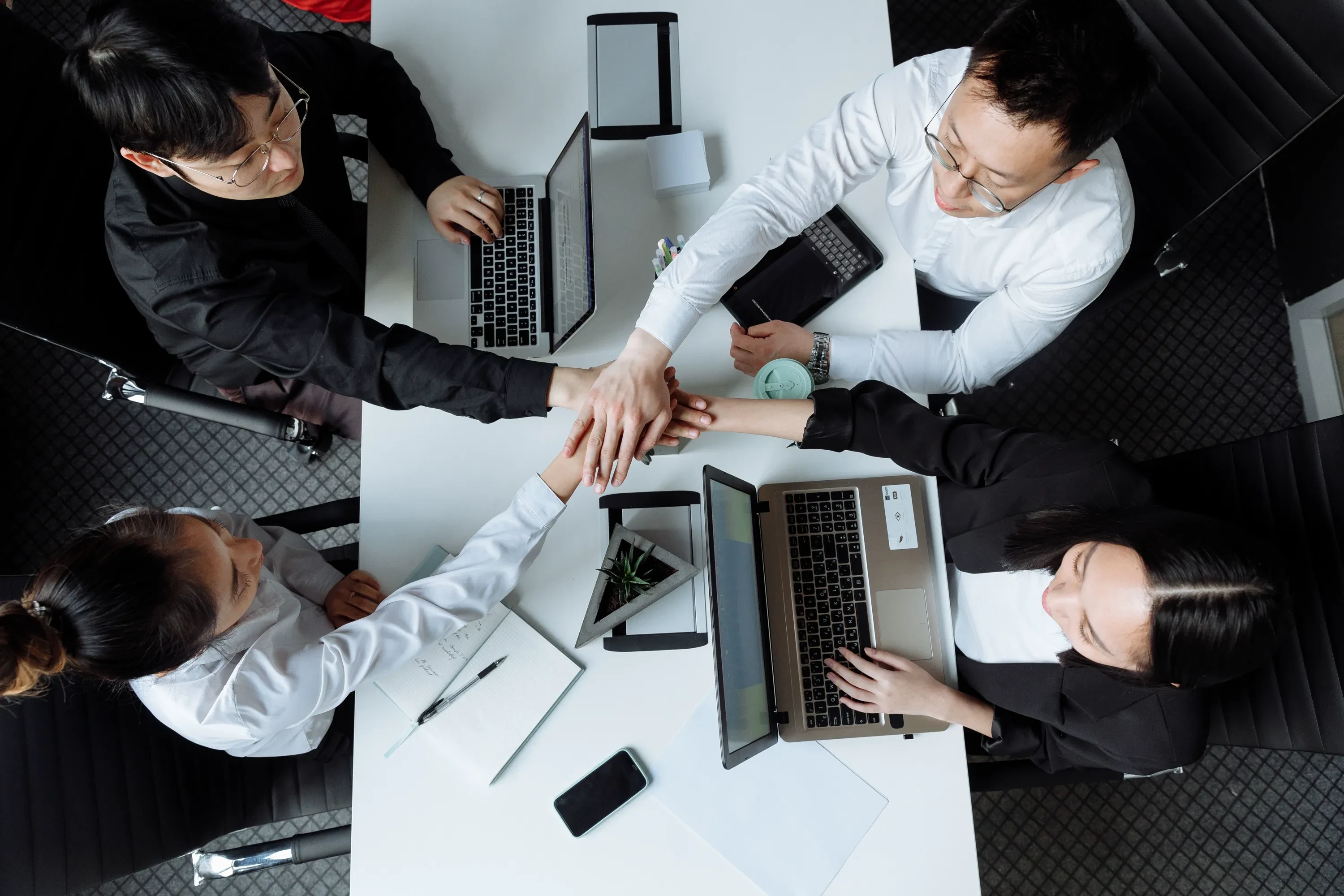 Overhead view of colleagues around a meeting table, their hands joined together beside open laptops